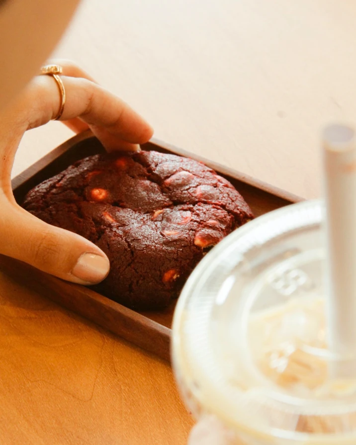 Mão pegando um cookie red velvet numa bandeja, ao lado de uma bebida gelada
